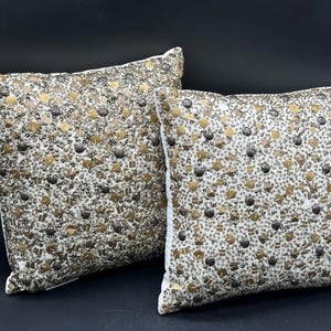 May include: Two square throw cushions featuring a dense, intricate surface covered in metallic gold and silver beads, sequins, and small decorative studs. The embellishments create a textured, reflective pattern across the entire front face.