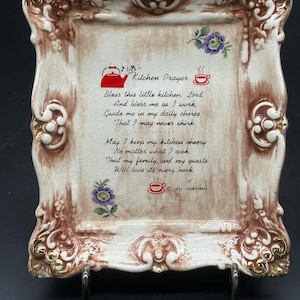 Op de afbeelding: Een rechthoekige keramische plaquette met een decoratieve bruine en gouden reliëflijst. Het midden bevat de tekst Kitchen Prayer, een gedicht, een rode theepot en twee illustraties van koffiekopjes. Kleine paarse bloemen sieren de hoeken.