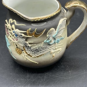 May include: A ceramic creamer pitcher featuring a raised, three-dimensional dragon motif in shades of white, blue, and gold against a dark gray and cream gradient background. The pitcher has a gold-trimmed rim and a gold-painted handle.