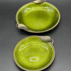 May include: Two round green dishes with metallic rims and leaf-shaped accents. The larger dish features a radial pattern, while the smaller dish has a mottled texture. Both items are finished with a twisted rope-style border and metal leaves.