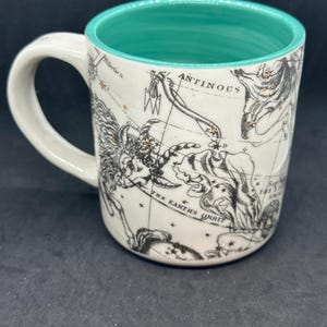 Puede incluir: Taza de cer&aacute;mica blanca con interior turquesa y asa. La taza presenta un dise&ntilde;o de mapa celeste negro y dorado con constelaciones y el texto "ANTINOUS" y "THE EARTH'S ORBIT".