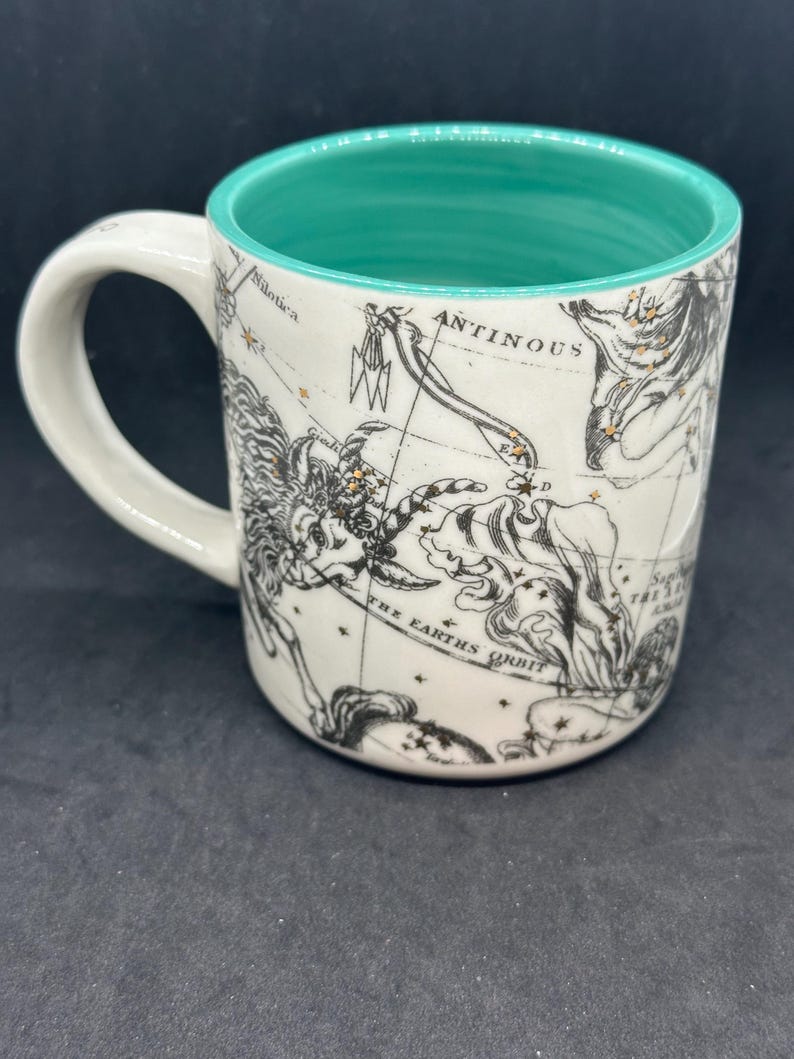 Puede incluir: Taza de cer&aacute;mica blanca con interior turquesa y asa. La taza presenta un dise&ntilde;o de mapa celeste negro y dorado con constelaciones y el texto "ANTINOUS" y "THE EARTH'S ORBIT".