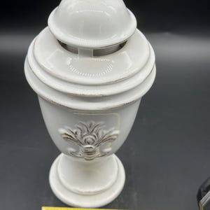 以下が含まれることがあります： 取り外し可能な蓋が付いた白いセラミック製の装飾用骨壺で、使い古された茶色のディテールが施された浮き彫りのフルール・ド・リスのエンブレムが特徴です。底面に置かれた黄色いメジャーは、幅が約11.5cmであることを示しています。