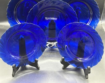 May include: A set of five cobalt blue glass plates featuring a swirling, scalloped edge design. The plates are arranged in a tiered formation, with two plates positioned on black stands in the foreground and three plates displayed behind them.