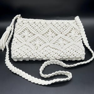 May include: An off-white macrame crossbody bag featuring a repeating diamond pattern with central star-like knots. The bag includes a braided shoulder strap and a decorative tassel attached to the side, all crafted from thick, textured cord.