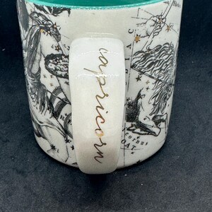 Puede incluir: Taza de cer&aacute;mica blanca con interior turquesa y asa. La taza presenta un dise&ntilde;o de constelaci&oacute;n en negro y dorado con la palabra "Capricornio" en escritura dorada en el asa. La taza tiene una est&eacute;tica vintage.