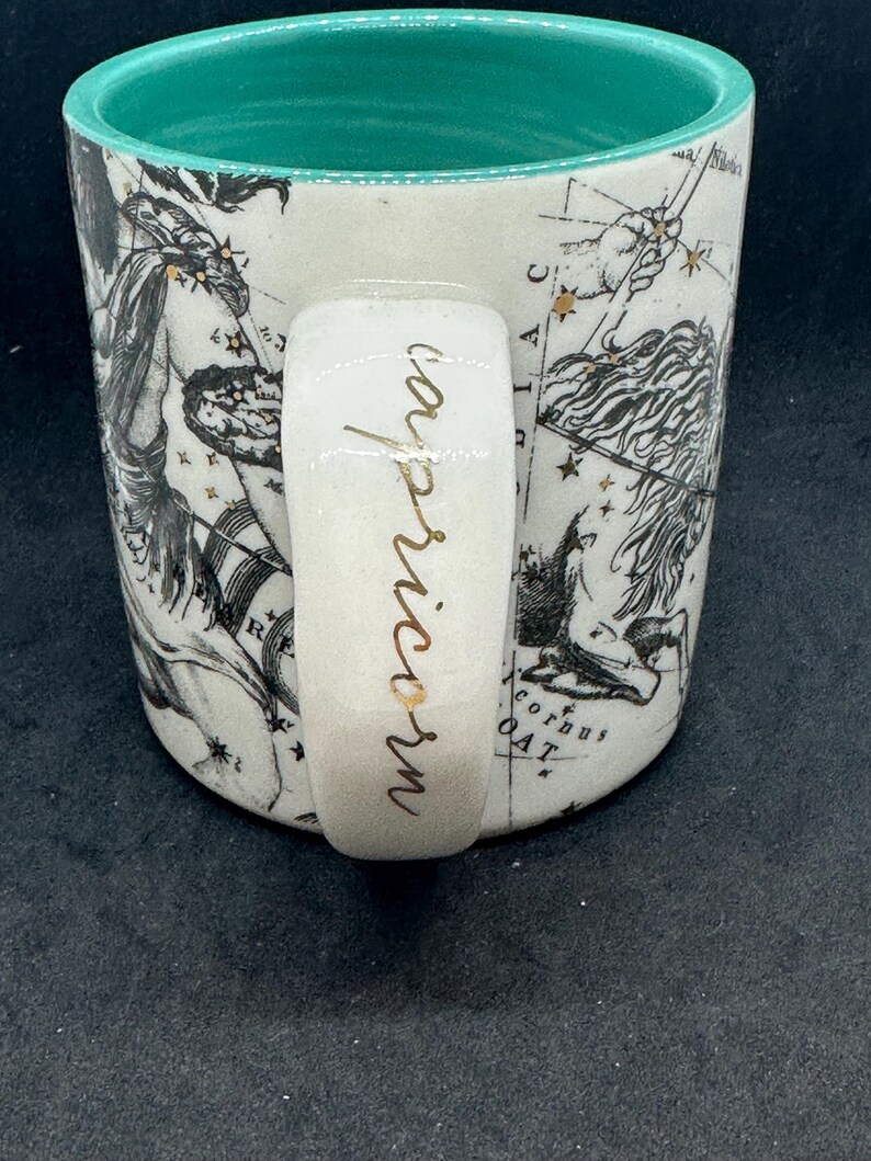 Puede incluir: Taza de cer&aacute;mica blanca con interior turquesa y asa. La taza presenta un dise&ntilde;o de constelaci&oacute;n en negro y dorado con la palabra "Capricornio" en escritura dorada en el asa. La taza tiene una est&eacute;tica vintage.