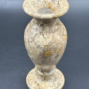 May include: A small, polished stone vase featuring a mottled, earthy pattern of beige, tan, and dark brown swirls. The vessel has a wide, flared rim, a rounded body, and a circular pedestal base, showcasing a natural, variegated mineral texture.