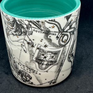Puede incluir: Un vaso de cer&aacute;mica blanco con interior turquesa. El exterior presenta un dise&ntilde;o de mapa vintage en negro y dorado con elementos celestes y n&aacute;uticos. El vaso tiene forma cil&iacute;ndrica y un acabado liso y brillante.