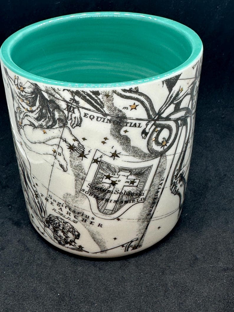 Puede incluir: Un vaso de cer&aacute;mica blanco con interior turquesa. El exterior presenta un dise&ntilde;o de mapa vintage en negro y dorado con elementos celestes y n&aacute;uticos. El vaso tiene forma cil&iacute;ndrica y un acabado liso y brillante.