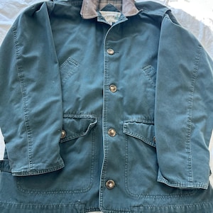 May include: A teal-colored jacket with a brown suede collar and four large gold buttons. The jacket has two large front pockets and a buttoned flap on each shoulder. The jacket is made of a durable material.
