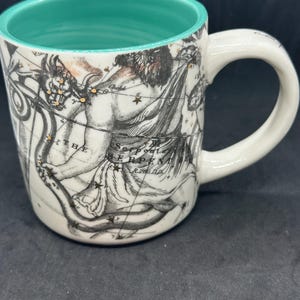 Puede incluir: Taza de cer&aacute;mica blanca con interior turquesa y asa. La taza presenta una detallada ilustraci&oacute;n en blanco y negro de una figura mitol&oacute;gica y constelaciones. El texto "THE SERPENT" es visible en la taza.