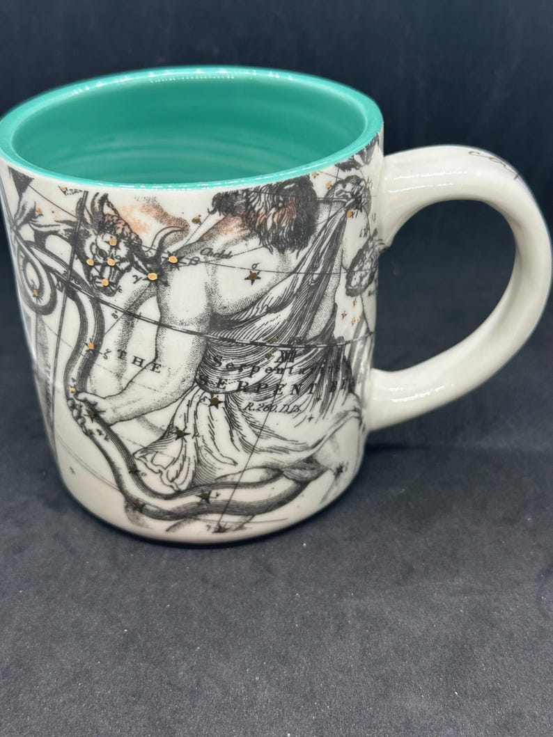 Puede incluir: Taza de cer&aacute;mica blanca con interior turquesa y asa. La taza presenta una detallada ilustraci&oacute;n en blanco y negro de una figura mitol&oacute;gica y constelaciones. El texto "THE SERPENT" es visible en la taza.
