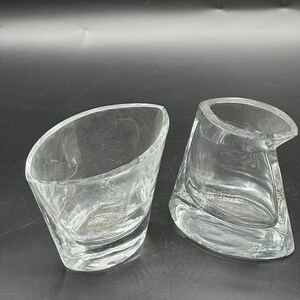 May include: Two clear glass vessels with asymmetrical, slanted rims and thick bases. One vessel is open and bowl-shaped, while the other features a distinct spout-like opening on one side, both showcasing smooth, transparent glass construction.