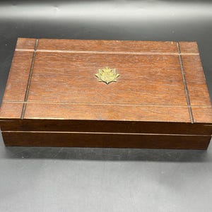May include: A rectangular dark brown wooden box with a smooth finish and a hinged lid. The top surface features a decorative border of incised lines and a centered, small, metallic gold-toned emblem shaped like a shield with intricate detailing.