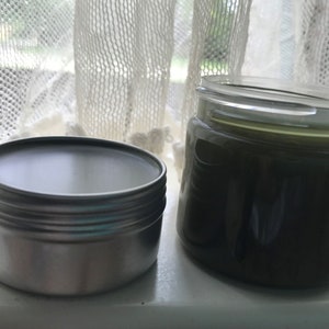 Pu&ograve; includere: Due contenitori di unguento alle erbe verde scuro, uno in una scatola di latta argentata e l'altro in un barattolo di vetro trasparente.