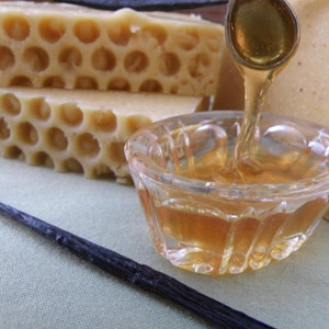May include: A small glass bowl filled with honey, with a spoon dripping honey into the bowl. There are two pieces of honeycomb soap and a vanilla bean on a green surface.