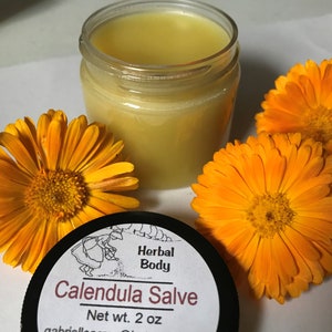 K&ouml;nnte beinhalten: Ein kleines Glasgef&auml;&szlig; gef&uuml;llt mit einer gelben Salbe, beschriftet mit "Calendula Salve" mit dem Text "Herbal Body" und "Net wt. 2 oz". Drei orangefarbene Ringelblumen umgeben das Gef&auml;&szlig;.