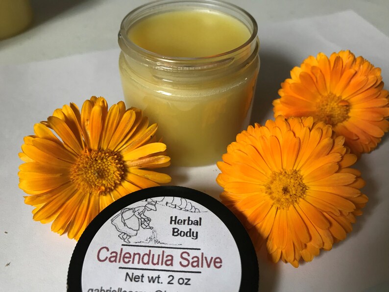 K&ouml;nnte beinhalten: Ein kleines Glasgef&auml;&szlig; gef&uuml;llt mit einer gelben Salbe, beschriftet mit "Calendula Salve" mit dem Text "Herbal Body" und "Net wt. 2 oz". Drei orangefarbene Ringelblumen umgeben das Gef&auml;&szlig;.