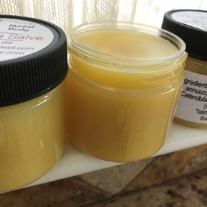 K&ouml;nnte beinhalten: Zwei Glasbeh&auml;lter gef&uuml;llt mit einer gelben Salbe. Die Beh&auml;lter haben schwarze Deckel und Etiketten mit dem Text "Herbal Body Calendula Salve", "net wt. 2 oz", "es@hotmail.com", "hend.etsy.com", "LA", und "gredients: "Sunflower (annuus) seed oil, Calendula officinalis), (Cera alba) *certified organic Batch # 1361".