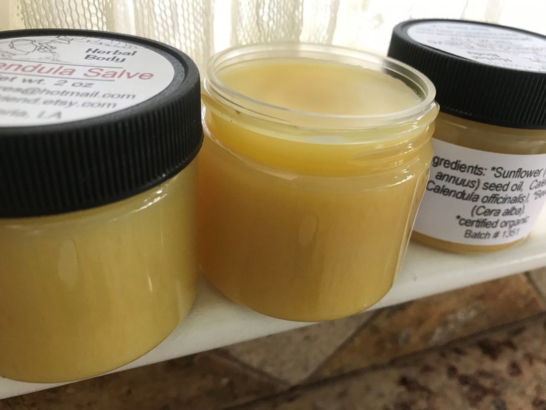 K&ouml;nnte beinhalten: Zwei Glasbeh&auml;lter gef&uuml;llt mit einer gelben Salbe. Die Beh&auml;lter haben schwarze Deckel und Etiketten mit dem Text "Herbal Body Calendula Salve", "net wt. 2 oz", "es@hotmail.com", "hend.etsy.com", "LA", und "gredients: "Sunflower (annuus) seed oil, Calendula officinalis), (Cera alba) *certified organic Batch # 1361".