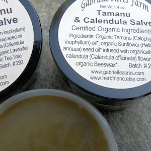 Pu&ograve; includere: Due piccoli barattoli di unguento Tamanu & Calendula, un unguento certificato biologico. I barattoli sono neri con etichette bianche che elencano gli ingredienti e il numero di lotto #292. L'unguento &egrave; di colore marrone chiaro.