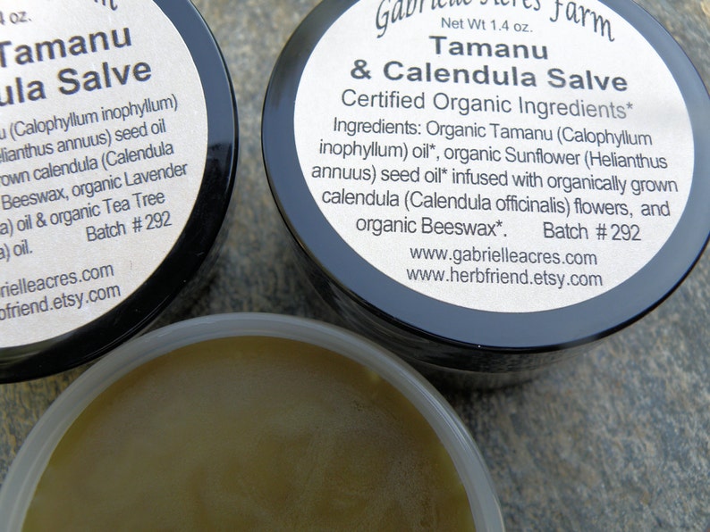 Pu&ograve; includere: Due piccoli barattoli di unguento Tamanu & Calendula, un unguento certificato biologico. I barattoli sono neri con etichette bianche che elencano gli ingredienti e il numero di lotto #292. L'unguento &egrave; di colore marrone chiaro.