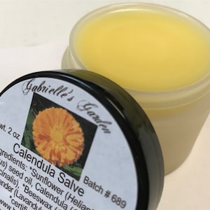 K&ouml;nnte beinhalten: Ein kleines, klares Glas mit schwarzem Deckel, das eine gelbe Salbe enth&auml;lt. Das Etikett auf dem Glas lautet "Calendula Salve" und "Gabrielle's Garden" mit einem Bild einer gelben Blume. Das Etikett enth&auml;lt auch den Text "Batch # 689" und "et wt. 2 oz".