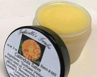May include: A small, clear jar with a black lid containing a yellow salve. The label on the jar reads "Calendula Salve" and "Gabrielle's Garden" with a picture of a yellow flower. The label also includes the text "Batch # 689" and "et wt. 2 oz".