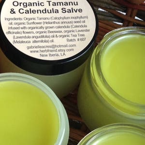 Pu&ograve; includere: Due barattoli di unguento biologico al tamanu e alla calendula di colore giallo. L'etichetta elenca gli ingredienti, tra cui olio di tamanu, olio di girasole, fiori di calendula, cera d'api, olio di lavanda e olio di tea tree. L'etichetta include anche il sito web e l'indirizzo email del venditore.