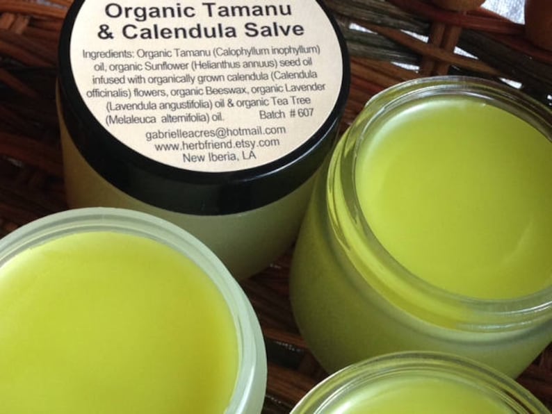 Pu&ograve; includere: Due barattoli di unguento biologico al tamanu e alla calendula di colore giallo. L'etichetta elenca gli ingredienti, tra cui olio di tamanu, olio di girasole, fiori di calendula, cera d'api, olio di lavanda e olio di tea tree. L'etichetta include anche il sito web e l'indirizzo email del venditore.