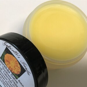 K&ouml;nnte beinhalten: Ein kleines, transparentes Glas gef&uuml;llt mit einer gelben Salbe. Das Glas hat einen schwarzen Deckel mit einem wei&szlig;en Etikett, auf dem "Calendula Salve" steht und die Zutaten aufgelistet sind: "Sunflower (Helianthus annuus) seed oil, Calendula (Calendula officinalis), Beeswax (Cera alba), Lavender (Lavandula angustifolia)". Das Etikett enth&auml;lt auch den Text "Gabrielle's Garden" und "Batch".