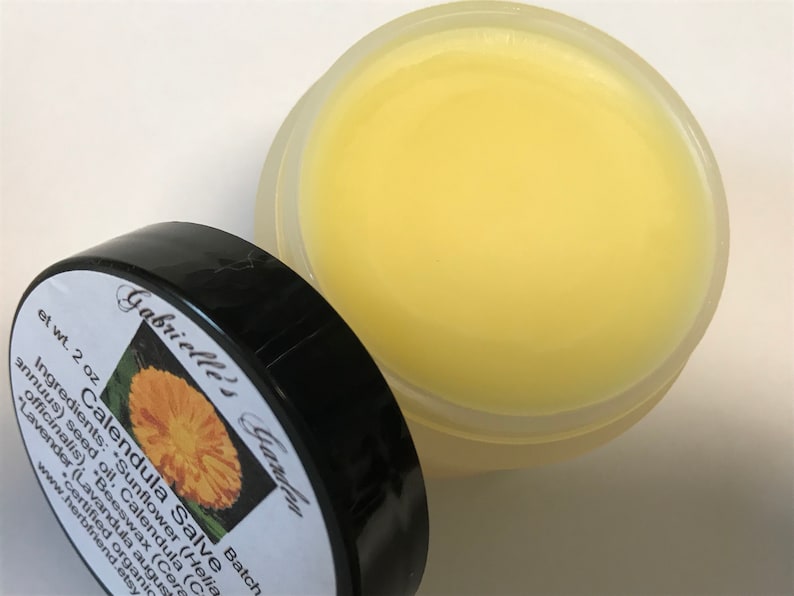 K&ouml;nnte beinhalten: Ein kleines, transparentes Glas gef&uuml;llt mit einer gelben Salbe. Das Glas hat einen schwarzen Deckel mit einem wei&szlig;en Etikett, auf dem "Calendula Salve" steht und die Zutaten aufgelistet sind: "Sunflower (Helianthus annuus) seed oil, Calendula (Calendula officinalis), Beeswax (Cera alba), Lavender (Lavandula angustifolia)". Das Etikett enth&auml;lt auch den Text "Gabrielle's Garden" und "Batch".