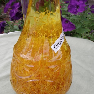 Pu&ograve; includere: Una bottiglia di vetro trasparente con una superficie testurizzata riempita di un liquido giallo e petali di fiori gialli. La bottiglia ha un'etichetta che dice "Organic".