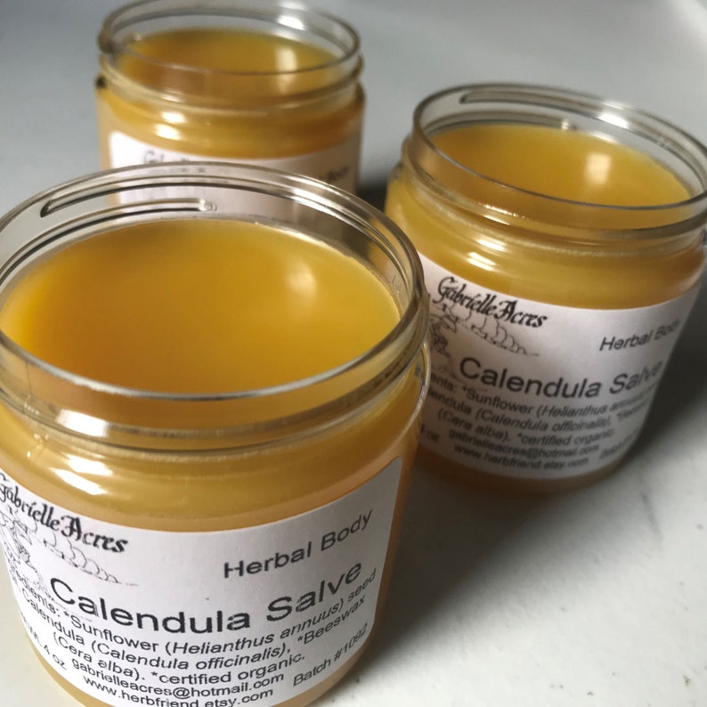 K&ouml;nnte beinhalten: Drei Glasbeh&auml;lter gef&uuml;llt mit einer gelben Salbe. Die Beh&auml;lter haben wei&szlig;e Etiketten mit dem Text "Gabrielle Acres" und "Calendula Salve" darauf gedruckt. Die Etiketten listen auch die Zutaten auf, darunter Sonnenblume, Calendula, Bienenwachs und zertifiziertes Bio-Cera Alba. Die Beh&auml;lter stehen auf einer wei&szlig;en Oberfl&auml;che.