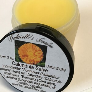 K&ouml;nnte beinhalten: Ein kleines, wei&szlig;es Glas mit schwarzem Deckel, beschriftet mit "Gabrielle's Garden", enth&auml;lt eine gelbe Calendula-Salbe. Das Etikett listet die Zutaten auf, darunter Sonnenblumenkern&ouml;l, Calendula, Bienenwachs und Lavendel&ouml;l. Das Glas ist mit "Calendula Salve" und "Batch # 689" beschriftet.