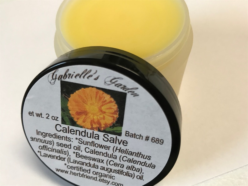 K&ouml;nnte beinhalten: Ein kleines, wei&szlig;es Glas mit schwarzem Deckel, beschriftet mit "Gabrielle's Garden", enth&auml;lt eine gelbe Calendula-Salbe. Das Etikett listet die Zutaten auf, darunter Sonnenblumenkern&ouml;l, Calendula, Bienenwachs und Lavendel&ouml;l. Das Glas ist mit "Calendula Salve" und "Batch # 689" beschriftet.