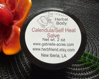 Könnte beinhalten: Ein runder schwarzer Behälter mit Calendula/Self Heal Salbe und einem weißen Etikett. Das Etikett zeigt eine Strichzeichnung einer Person, die Pflanzen gießt, den Text Herbal Body, Net wt. 2 oz, zwei Webadressen und den Ort New Iberia, LA.