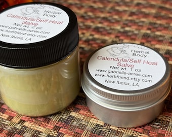 Könnte beinhalten: Zwei Behälter mit Calendula/Self Heal Salbe. Links ein durchsichtiger Tiegel mit schwarzem Deckel und 57g hellgelber Salbe. Rechts eine silberne Metalldose mit 28g Salbe. Beide haben weiße Etiketten mit schwarzer und roter Beschriftung.