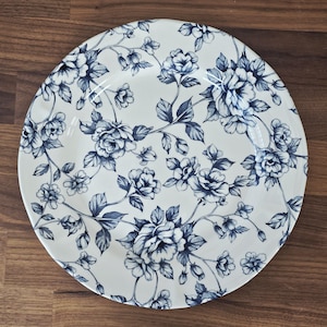 May include: A white ceramic plate with a blue floral pattern. The design features detailed blue roses and leaves, creating a classic and elegant aesthetic. The plate is round and sits on a wooden surface.