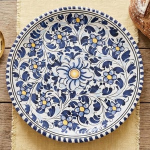 May include: A round ceramic plate featuring a hand-painted floral pattern in shades of dark blue and light blue with yellow centers. The design includes a large central flower surrounded by smaller blossoms and swirling leaf motifs.