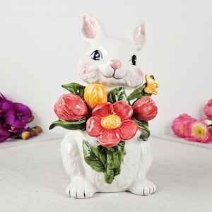 May include: A white ceramic bunny figurine holding a bouquet of flowers. The bunny has pink accents on its ears and cheeks, with a smiling face. The bouquet includes red, yellow, and green flowers, and a small yellow butterfly.