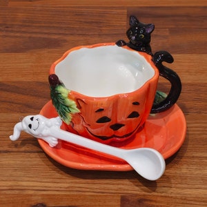 May include: A Halloween-themed ceramic teacup and saucer set. The orange teacup is shaped like a pumpkin with a black cat handle. A white ghost-shaped spoon rests on the orange saucer. The set is designed for Halloween.