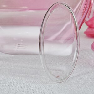 May include: Close-up of a clear, double-walled glass with a rounded oval shape. The glass is partially filled with a pink liquid, and the background is a soft pink. The glass sits on a white surface.
