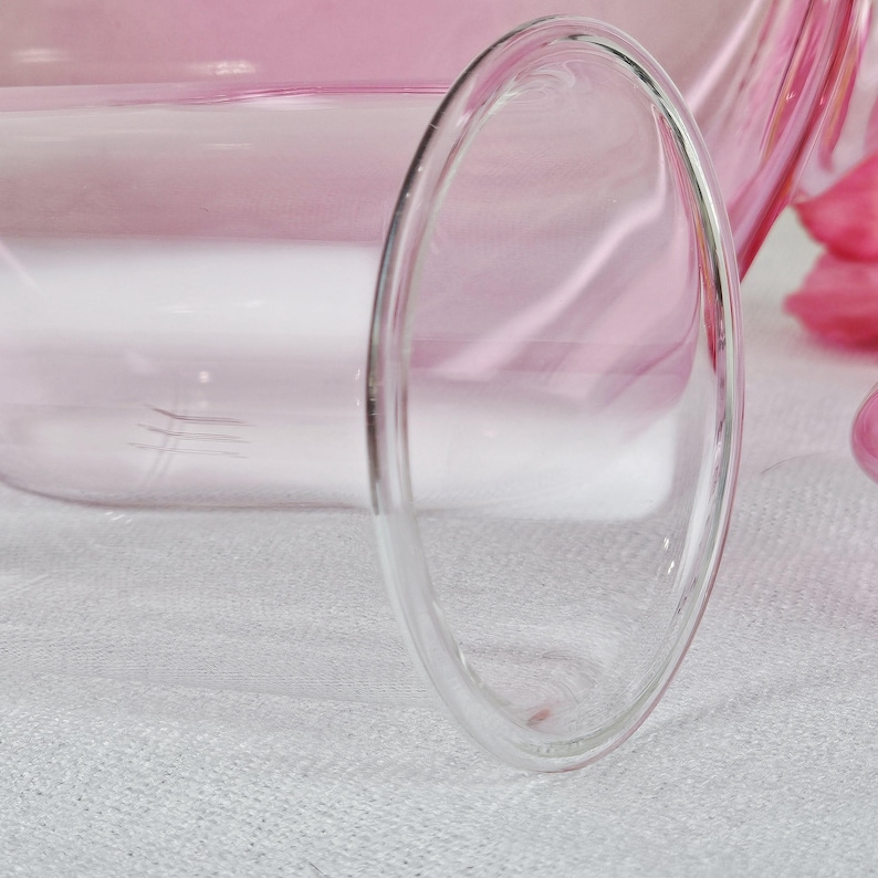 May include: Close-up of a clear, double-walled glass with a rounded oval shape. The glass is partially filled with a pink liquid, and the background is a soft pink. The glass sits on a white surface.