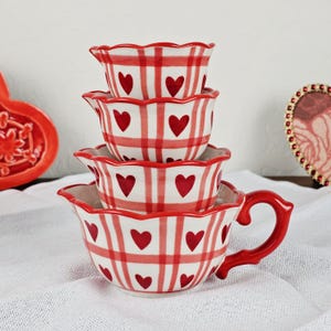 May include: A set of four ceramic measuring cups, featuring a red and white checked design with red heart motifs. The largest cup has a handle. The cups are stacked on a white surface.