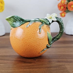 May include: A ceramic creamer shaped like a textured orange with a green branch handle, a green leaf spout, and a white citrus blossom detail. The surface features the characteristic dimpled skin of an orange in shades of bright orange and yellow.