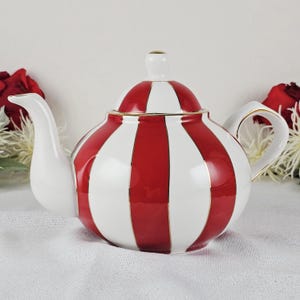 May include: A ceramic teapot with a red and white striped pattern. The teapot has a white spout, handle, and lid with a gold trim. The teapot is designed for serving tea.