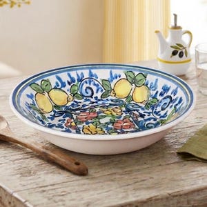 May include: A round ceramic bowl featuring a hand-painted pattern of yellow lemons with green leaves and blue floral accents. The bowl has a white interior and exterior with a blue rim. A wooden spoon rests nearby on the wooden surface.