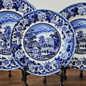 May include: A collection of blue and white decorative plates featuring a floral and landscape design. The plates are displayed on black metal stands. The plates have a detailed floral border and a central image of a bridge and building.
